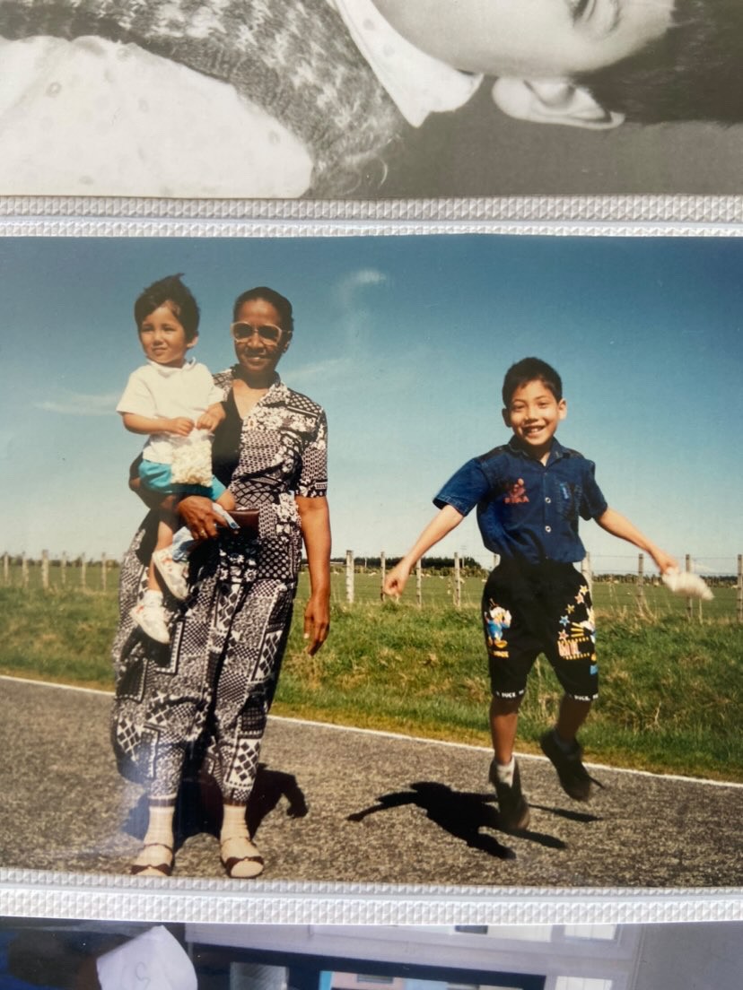 Woman in black and white patterned dress and round rimmed glasses holds a toddler in a white t-shirt, blue shorts, and white sneakers. An child is next to then jumping into the air wearing black shorts and a blue denim shirt. They are on a road in front of a fenced grass farmland.