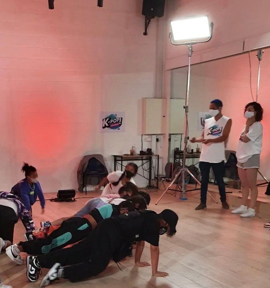 A group of dancers on the ground being watched over by two teachers in masks