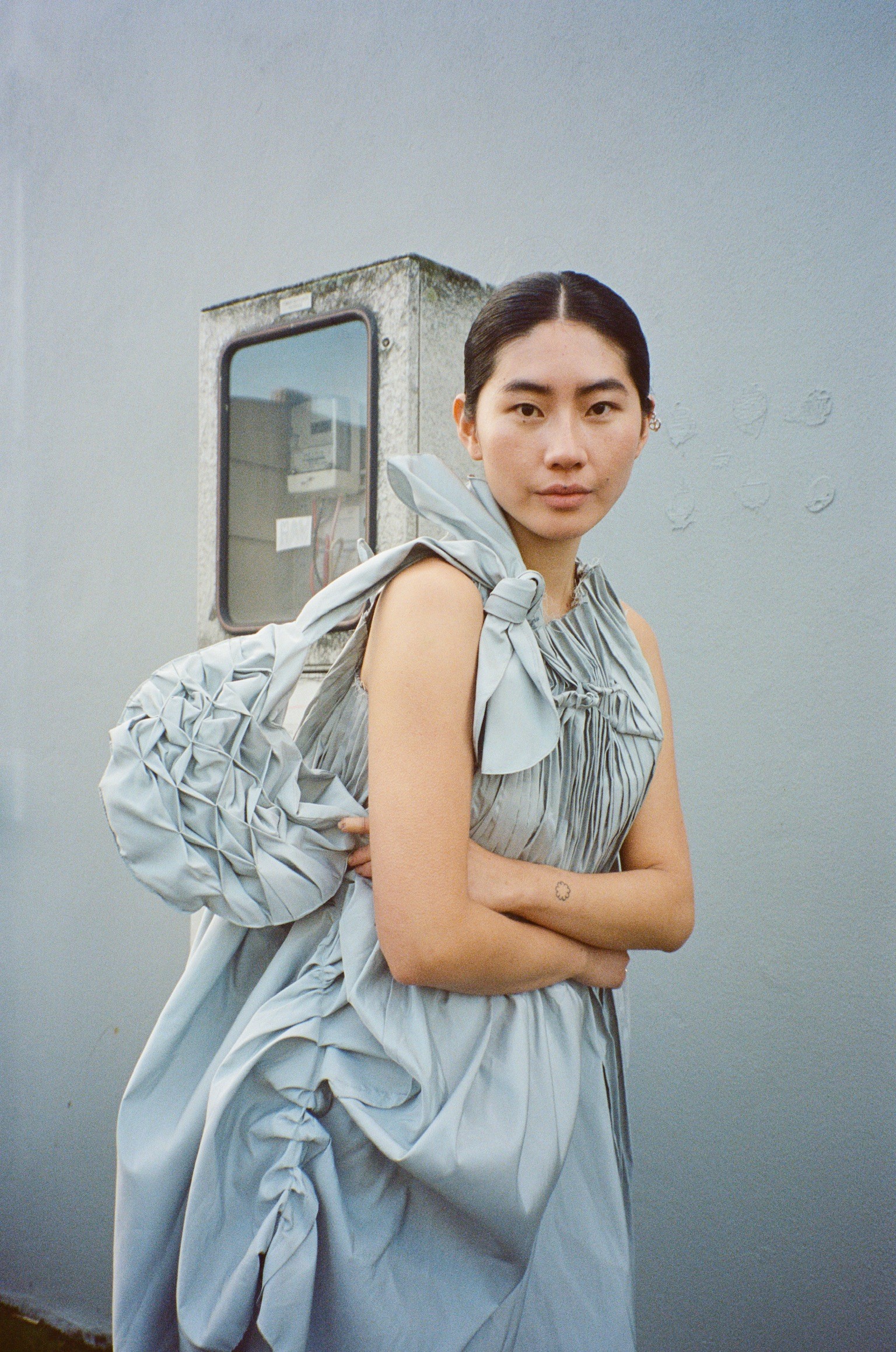 A model in a dusky blue dress with ruching and a matching bag crosses her arms and poses for the camera