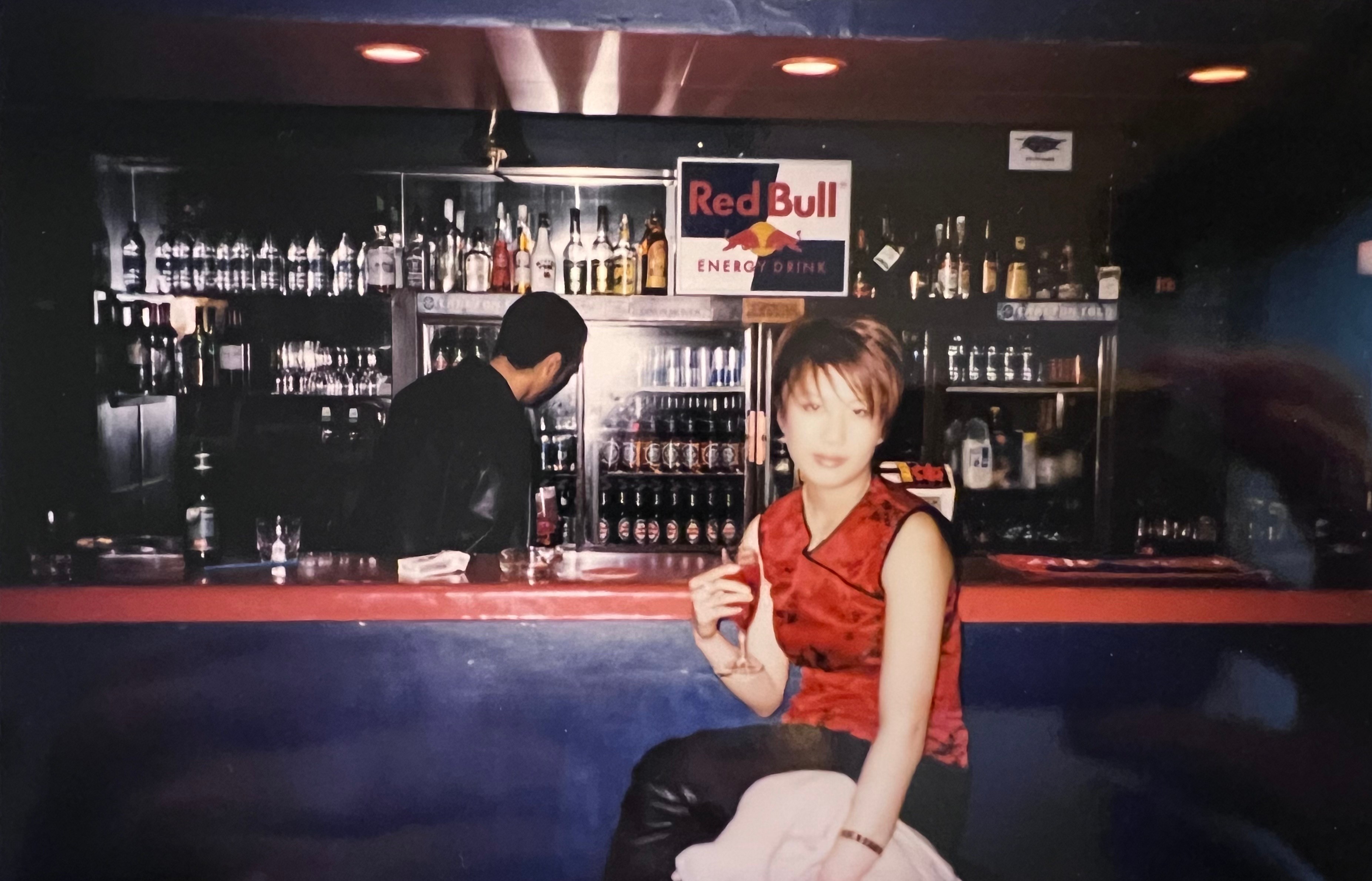 A woman in a red cheongsam sits with a glass of red wine at a bar. A man, with his back turned to the camera, stands behind the bar in front of a wall of liquor bottles and a Red Bull sign.