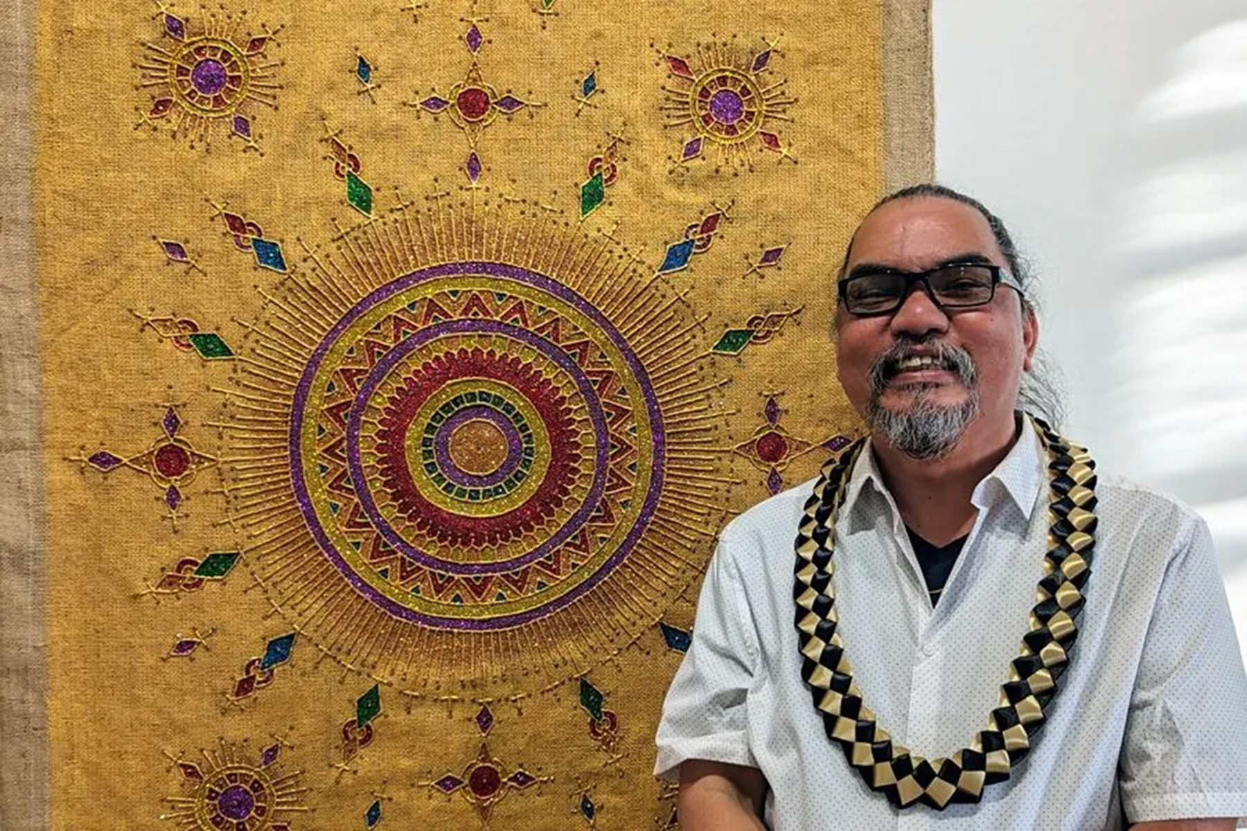 A smiling FIlipino wearing a woven black and white necklace standing in front of a stylised painting of a sun