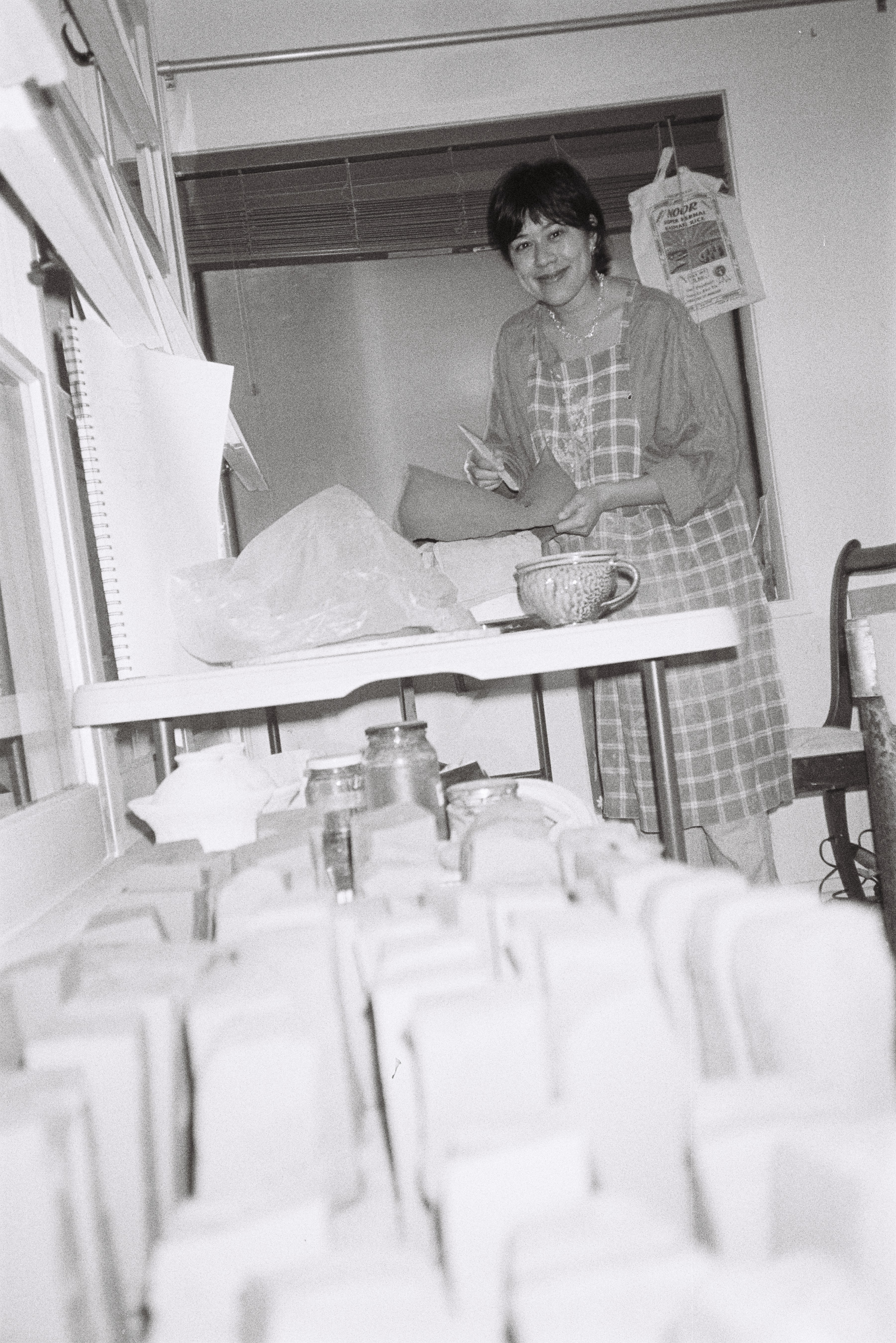 Black and white film low-angle photo of pottery with a woman in a plaid dress standing in the background, smiling