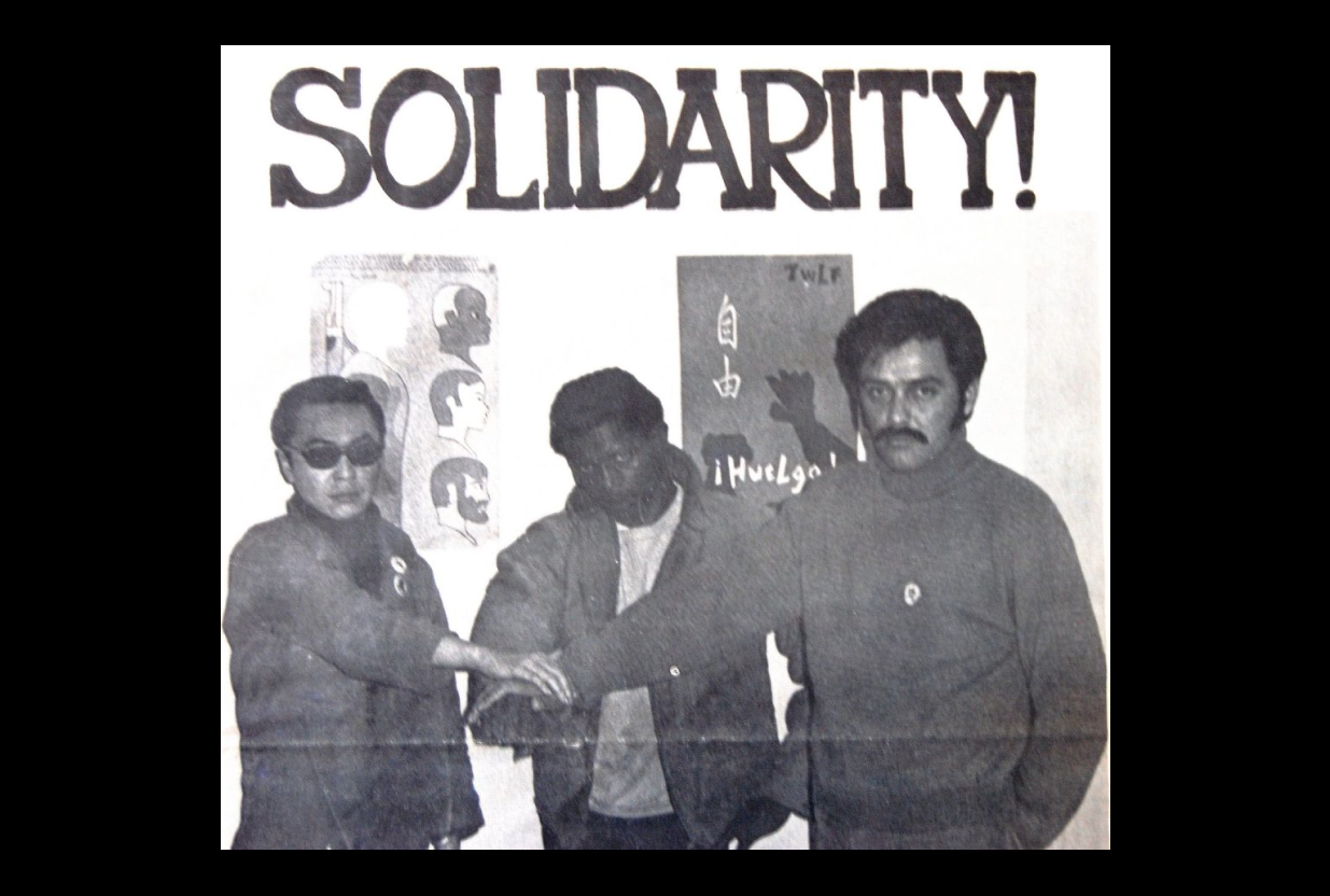 Three men with hands stacked in solidarity stand in front of posters.