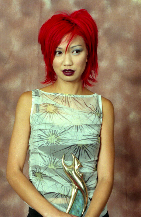A woman with a red bob and a sheer silver tank top holds an award
