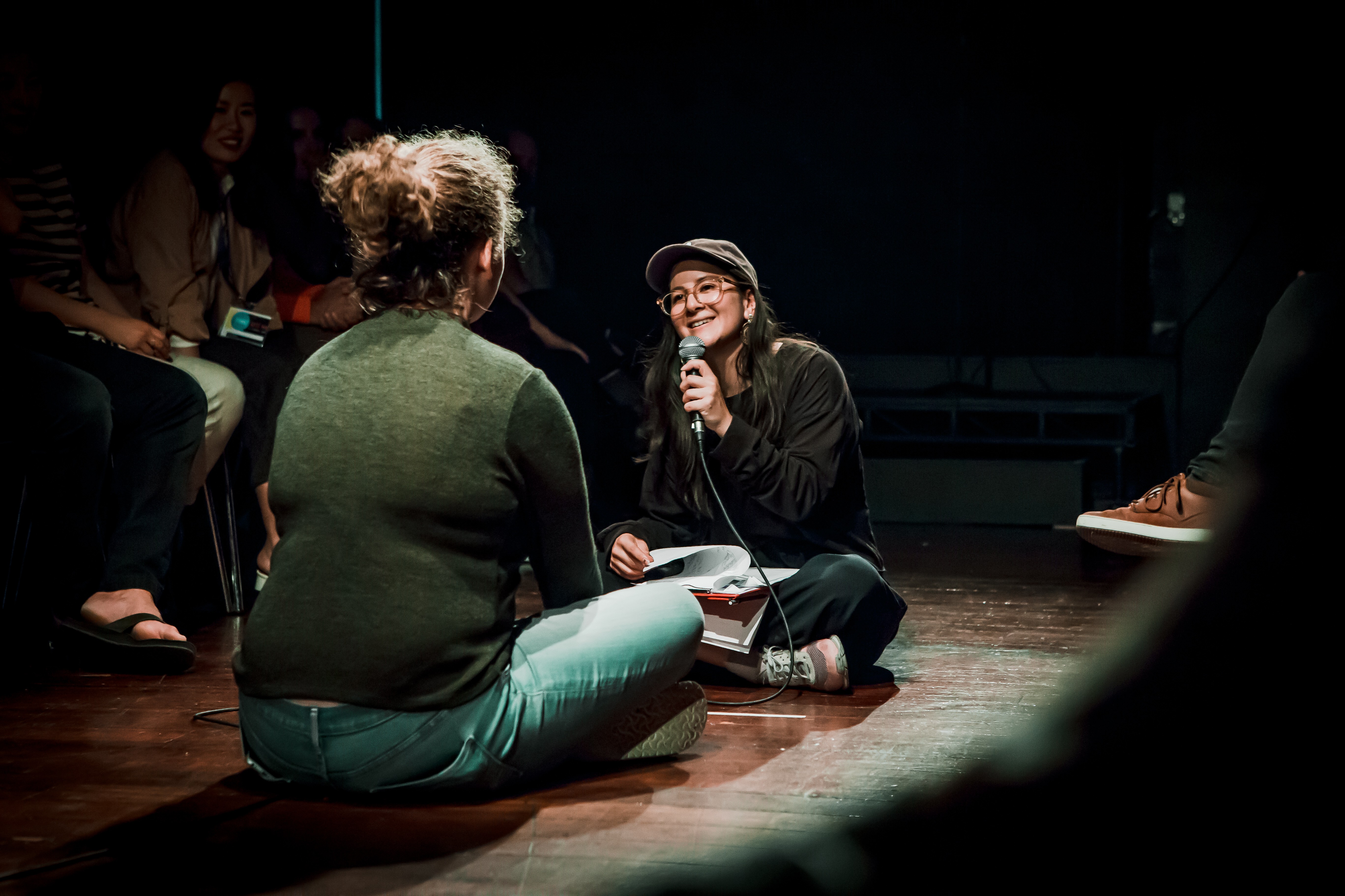 Two women sit cross-legged on the floor facing each other. One is holding a microphone interviewing the other