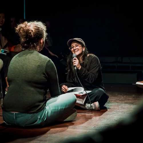 Two women sit cross-legged on the floor facing each other. One is holding a microphone interviewing the other