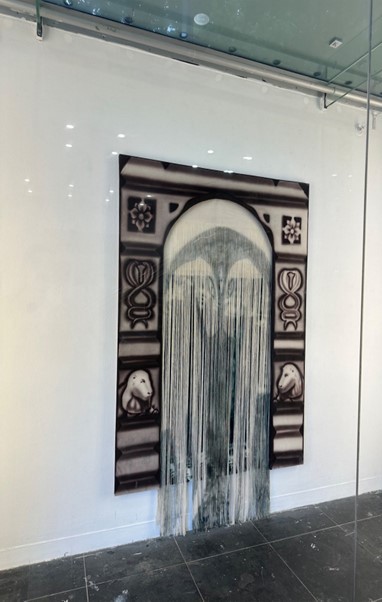 A painting hung on a white wall with frabric fringe detail