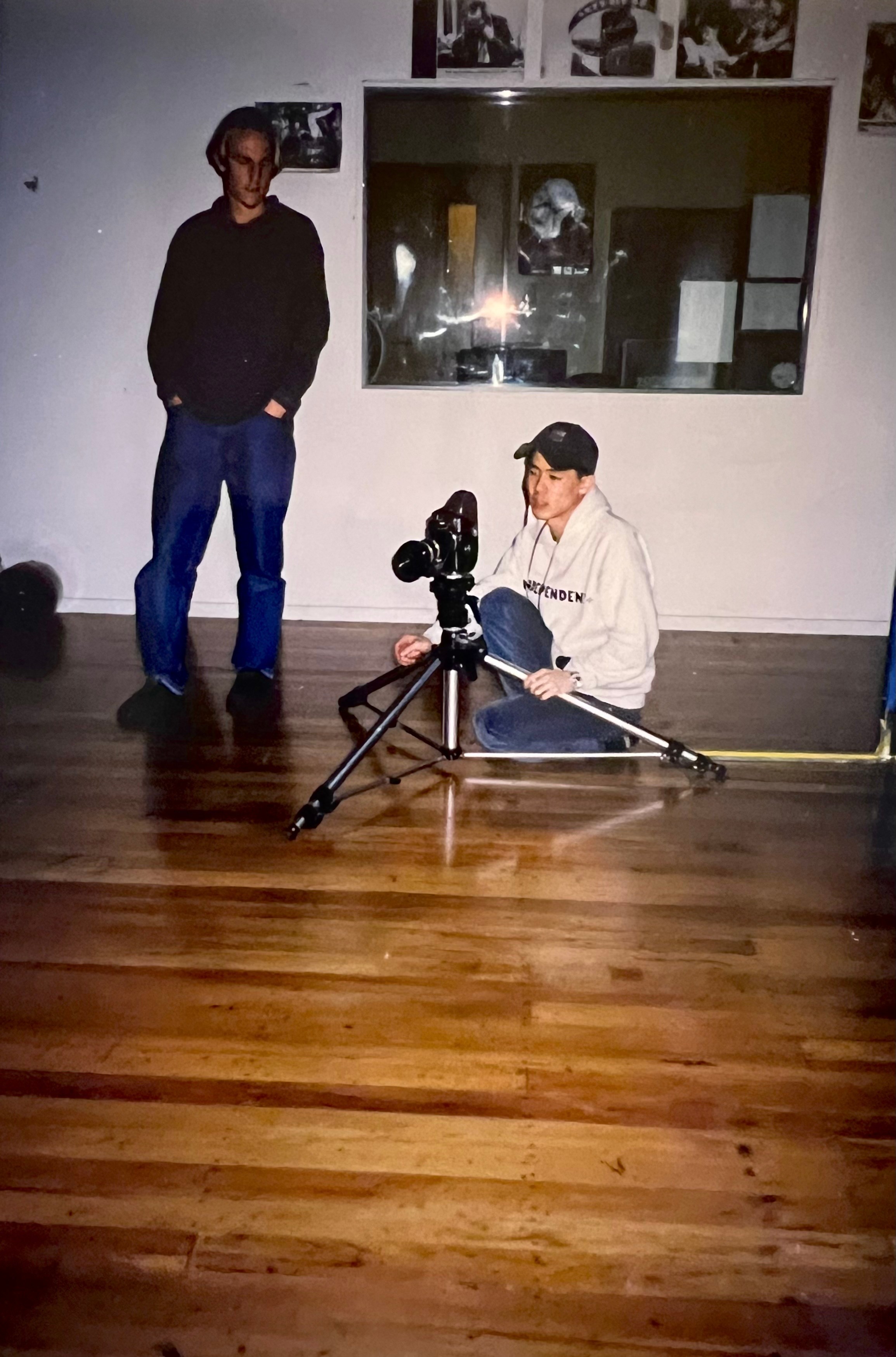 A man in blue jeans stands next to a man in a white hooded sweatshirt and black baseball cap, who is crouched next to a camera and tripod.