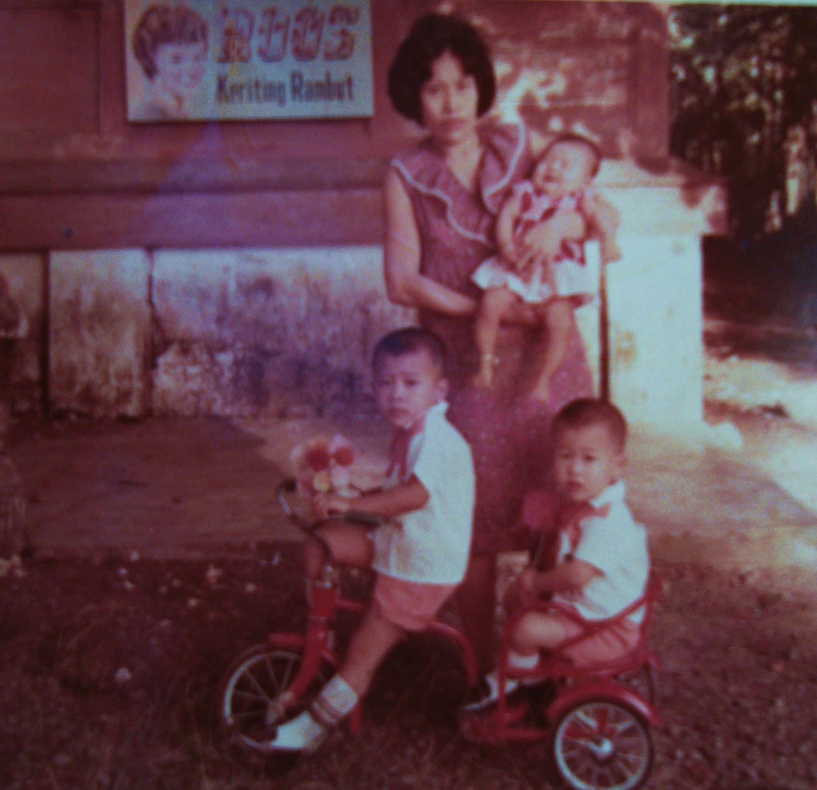 Photo of a woman standing outdoors in Indonesia with three children: a baby in her arms and two boys on a bike.