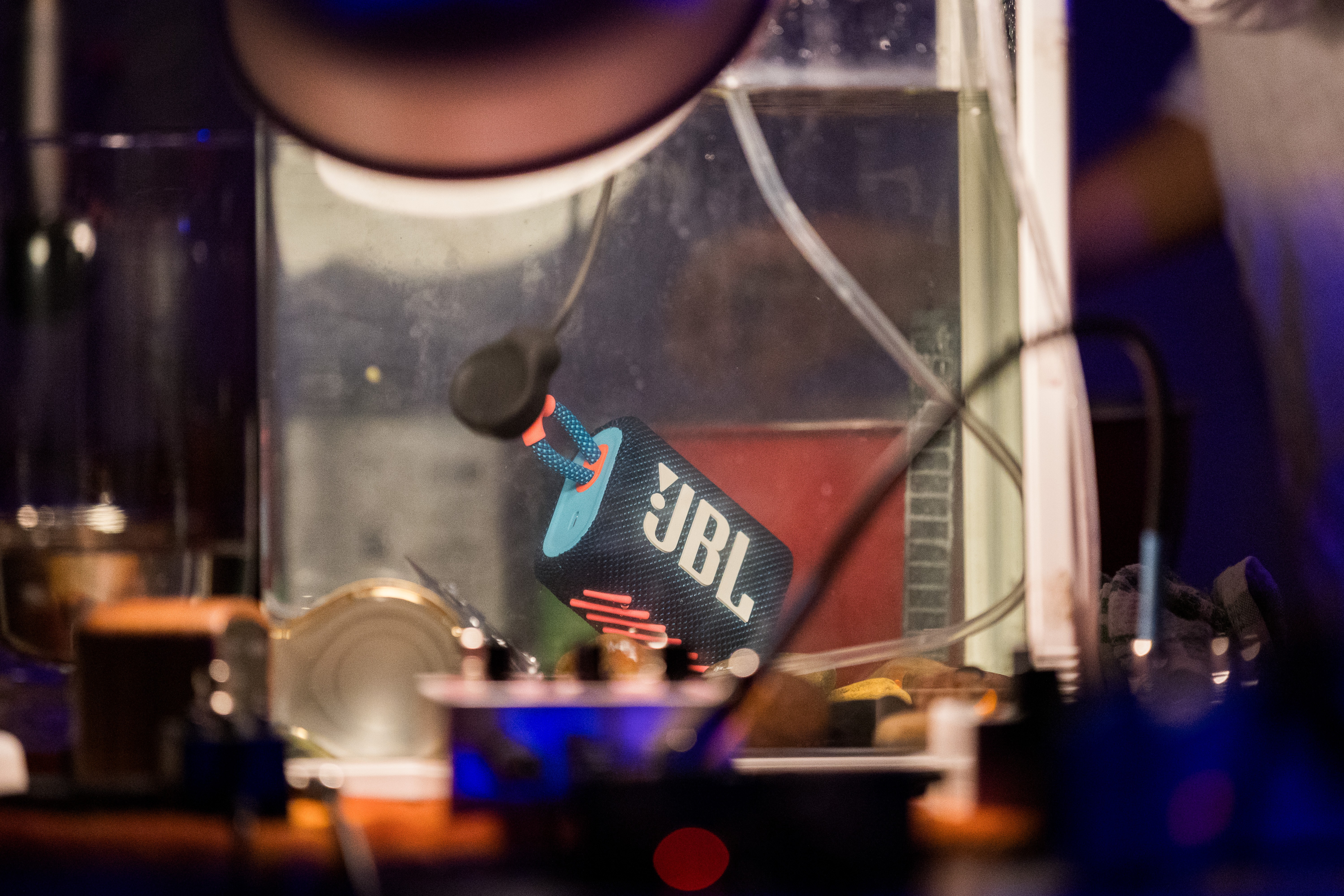 A speaker is submerged in a fishtank full of water. Cords and electronic equipment are tangled in the background of the image.
