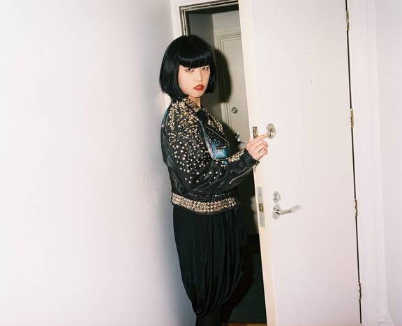 Dark haired woman wearing a studded leather jacket and red lipstick going through a door and looking back at the camera  