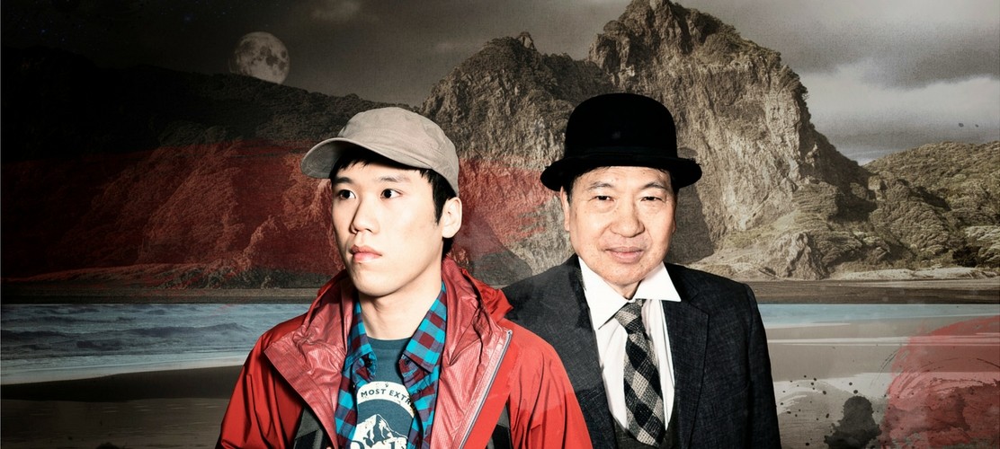 A young Chinese man in a red rain jacket and cap stands on a beach while a man in a suit stands behind him