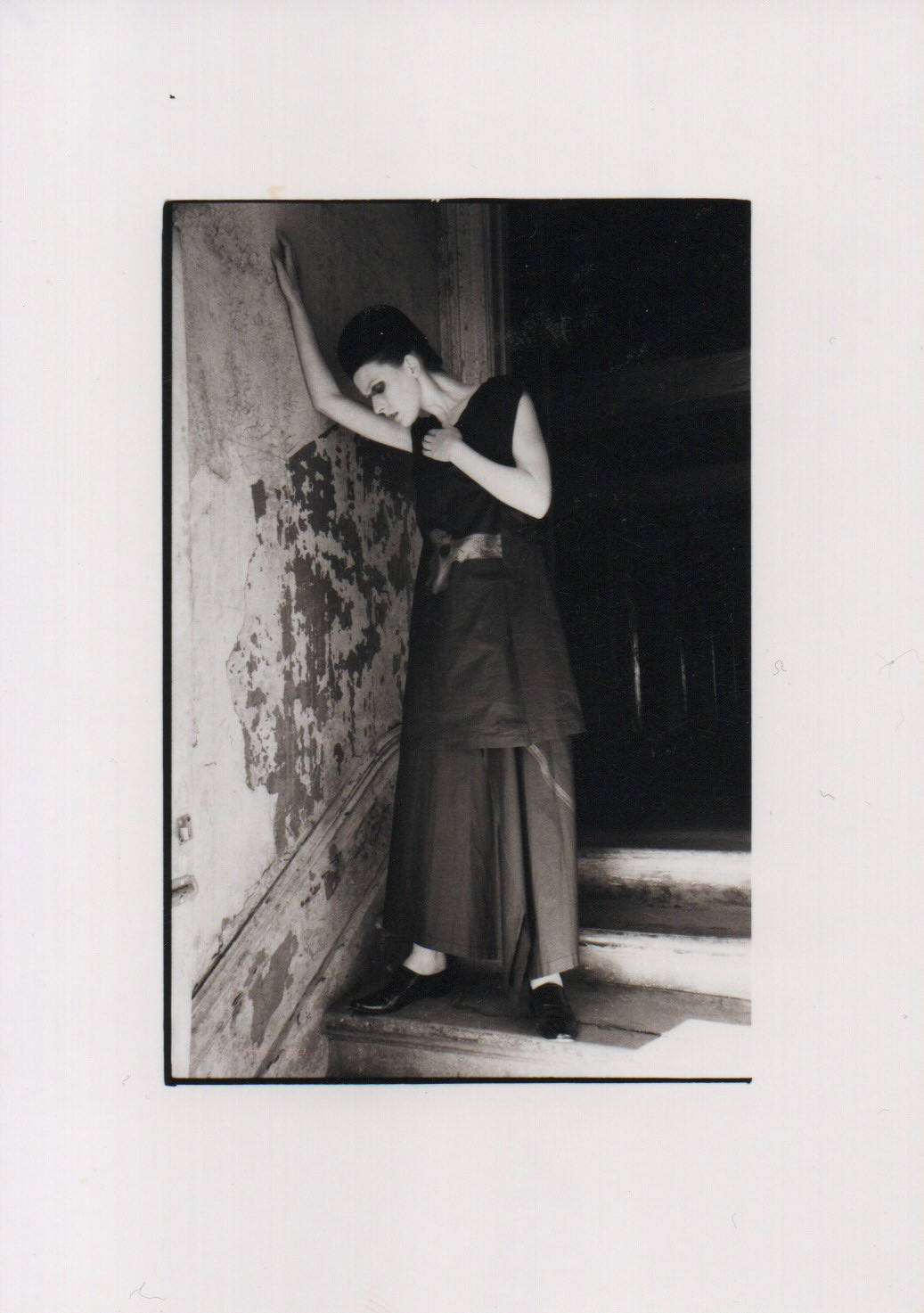 A black and white photo of a woman in a draped floor-length outfit leaning against a stone wall