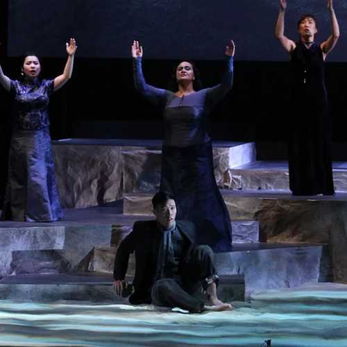 A stage with a chorus of seven women raising their hands to the sky. Three men stand in front of them, blue light washes over them