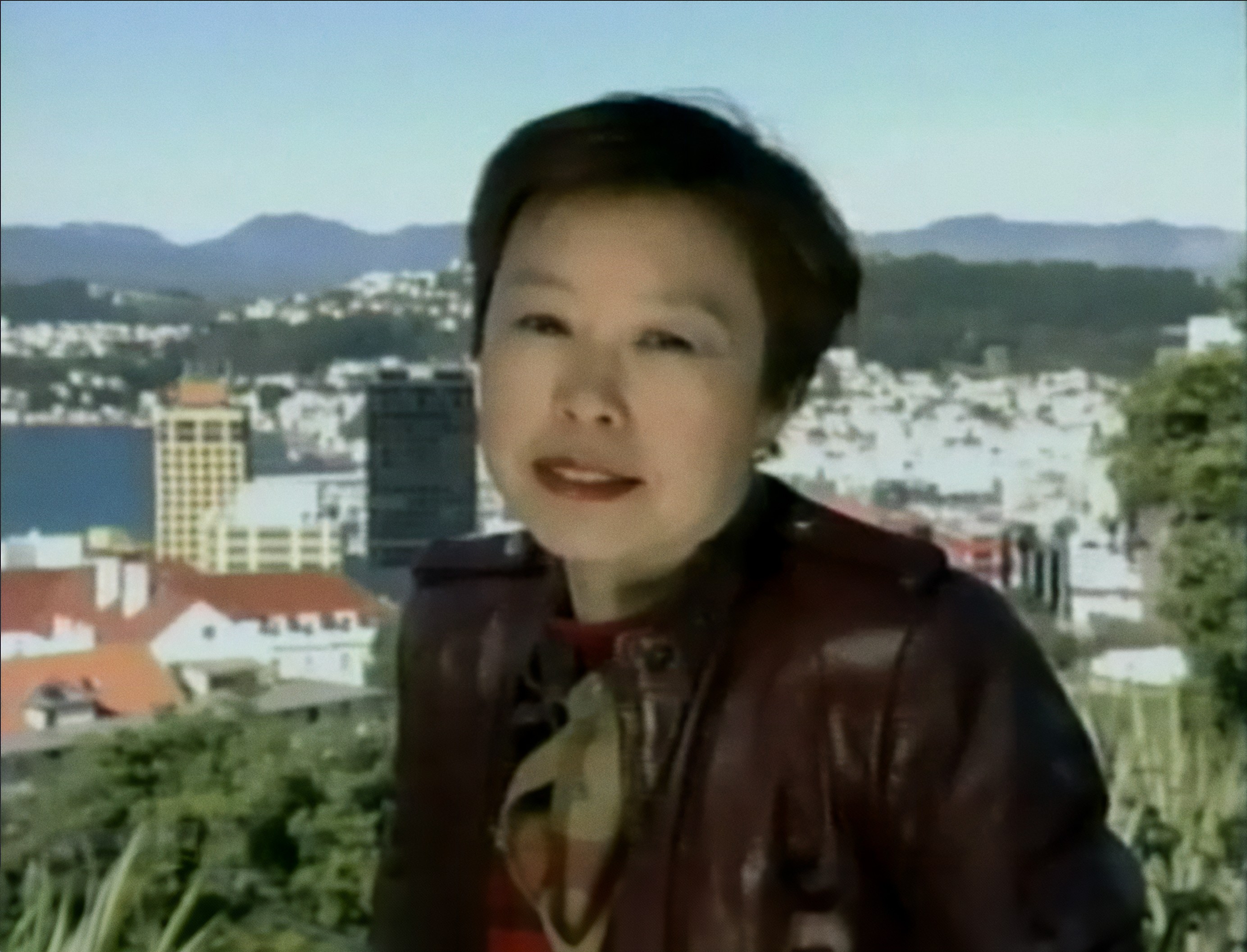 A woman in a maroon leather jacket stands outdoors with a cityscape and mountains in the background under a clear blue sky.