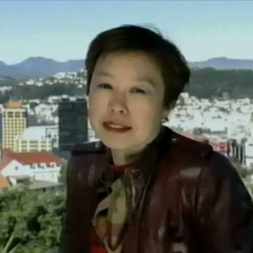 A woman in a maroon leather jacket stands outdoors with a cityscape and mountains in the background under a clear blue sky.