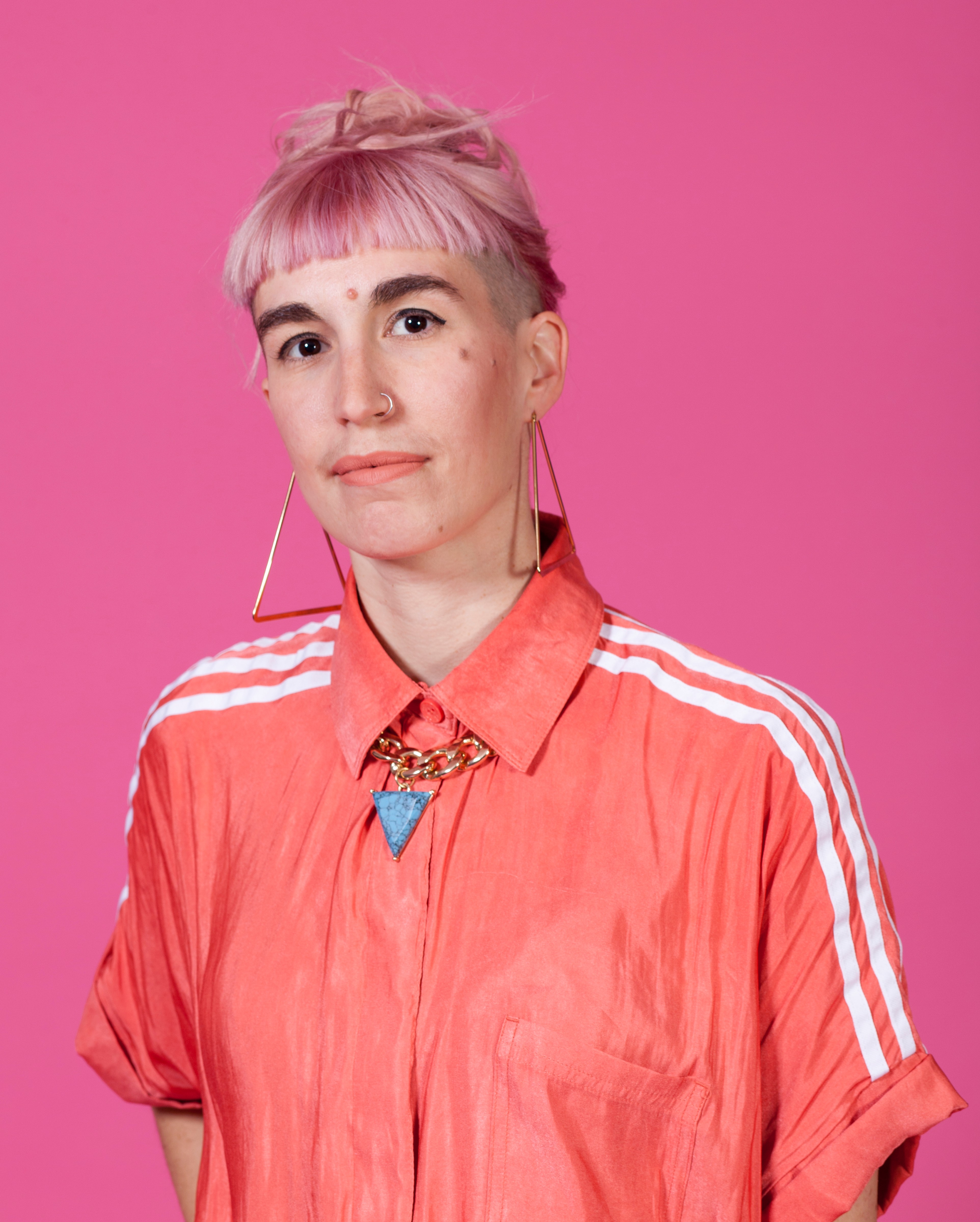 A woman with a short pink fringe and large triangle earrings wearing an apricot shirt with white stripes.
