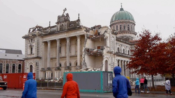 In a scene from the documentary ‘Moving’, three people in raincoats walk past an earthquake-damaged historical building in central Christchurch, New Zealand.