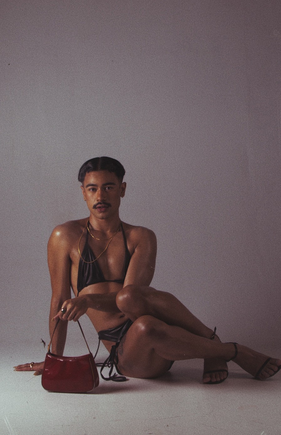 A gender bending model with a moustache and a masculine haircut wearing a two-piece bikini with sandals and long nails