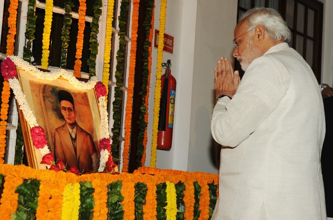 A portrait is surrounded by garlands. Modi stands with palms together facing the portrait.