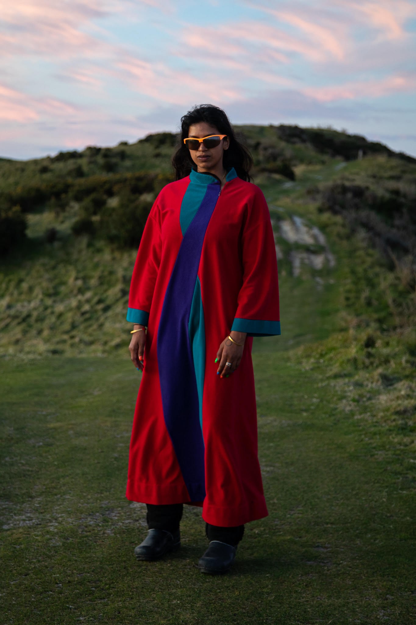 Photo of a person standing outside on a field with a hill behind them at dusk. They have long black hair and are wearing sunglasses with orange trim and a long loose-fitting red, teal, and purple dress