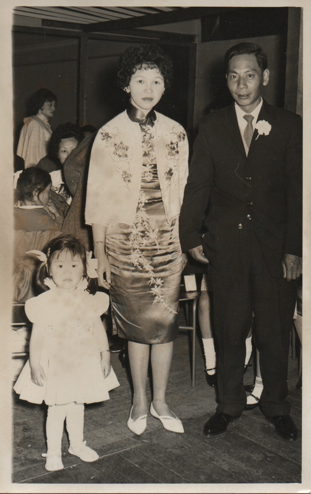 Black and white photo of a young baby in a white dress standing with her parents.