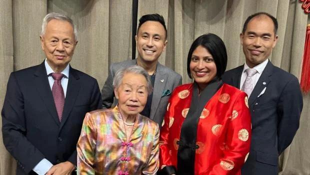 Five Asian people of different ages and genders, dressed formally, pose for a group photo at an event.