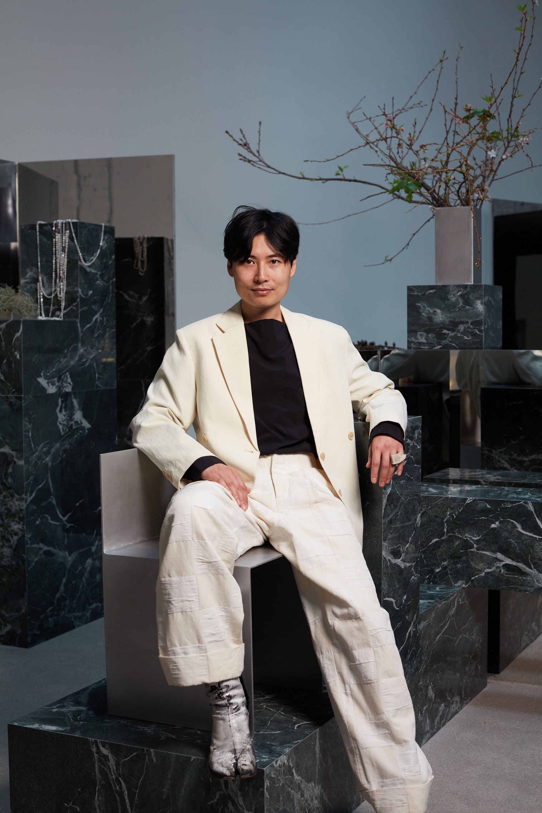 A man with black hair wearing a cream suit leans against dark green marble plinths