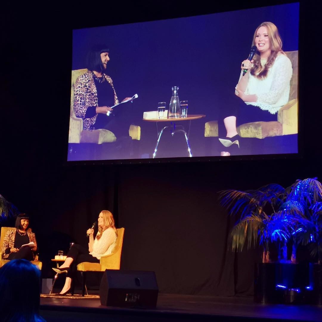 Two people seated and talking on a stage with a live screen projected above them