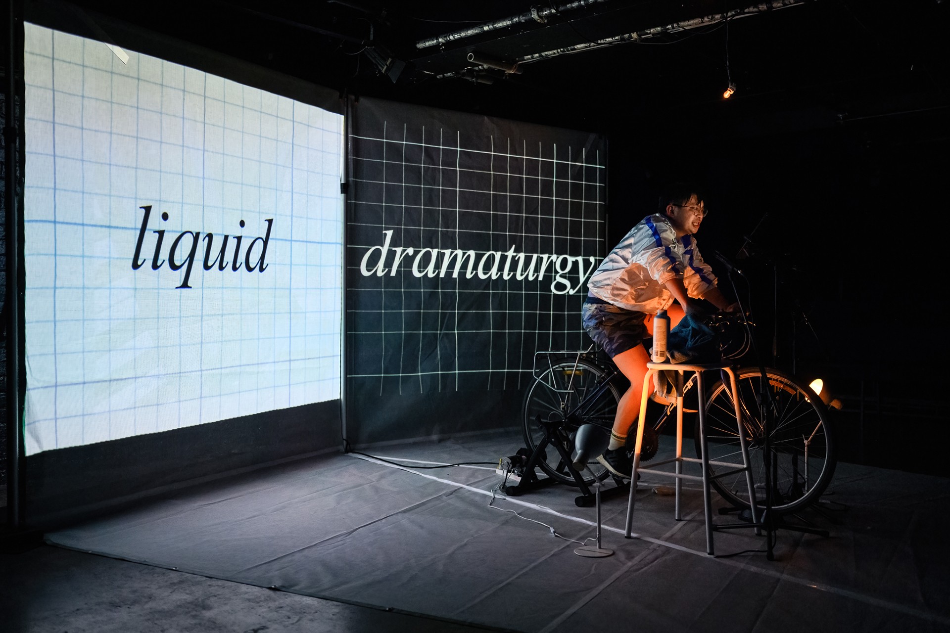 Nathan Joe on an exercycle on stage in front of a projection that reads 'liquid dramaturgy'.