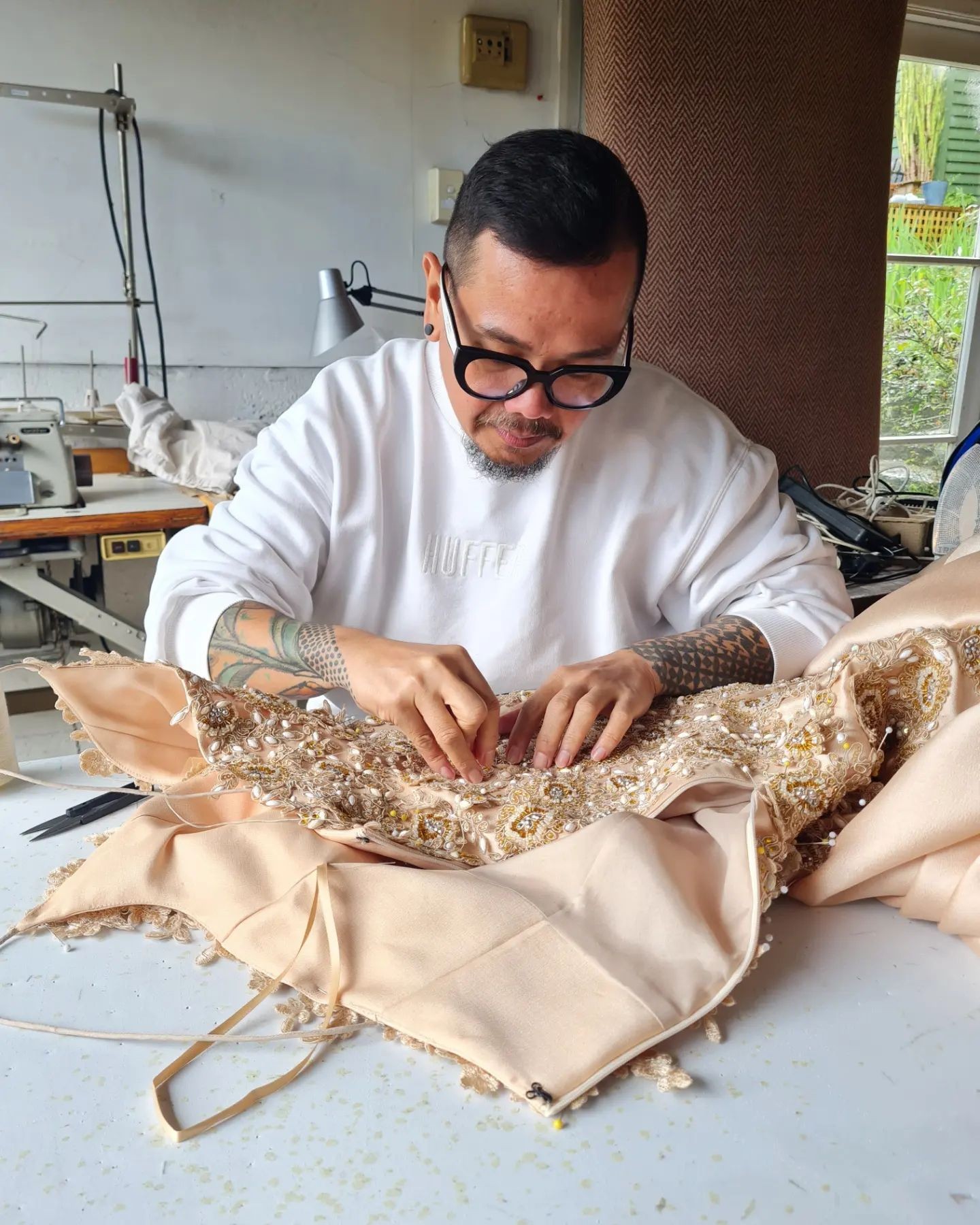 A Filipino man wearing horn-rimmed glasses and a white top is sewing beads into a cream-coloured gown