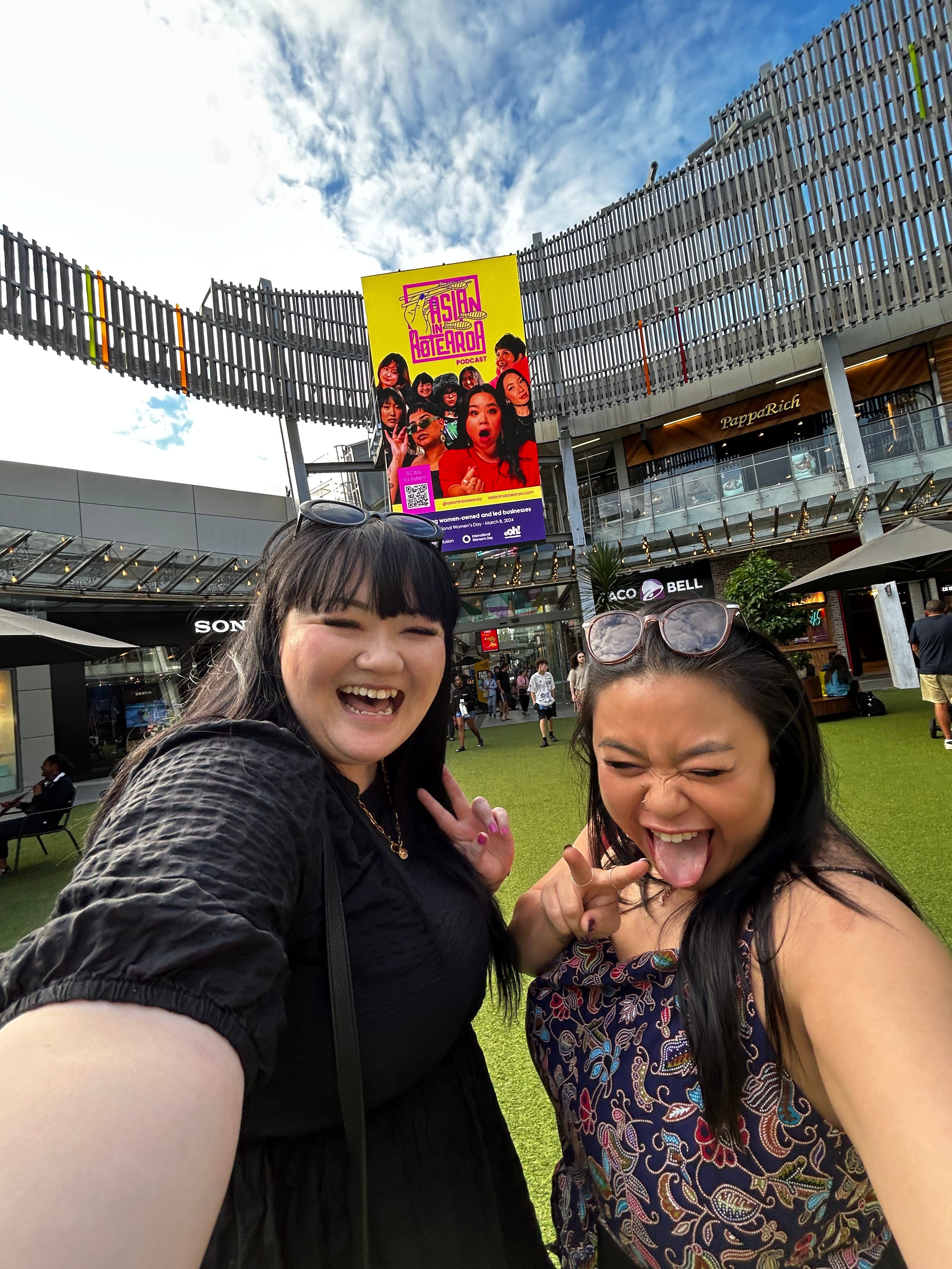 Two people taking a selfie with a billboard in the background with 'Asian in Aotearoa' on it