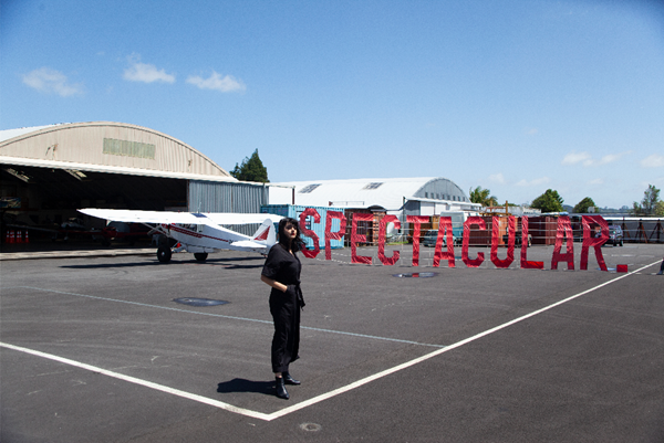 Photo outside of a plane hanger with a small white aeroplane in front of it that has a banner with the word "SPECTACULAR." attached to it. A person in black clothing with their hands in their pocket stands in the foreground.