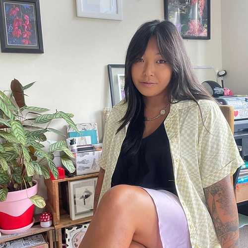A woman sitting in front of a wall with framed art work. She has long black hair and is wearing a black t-shirt, pale yellow shirt, and purple skirt.
