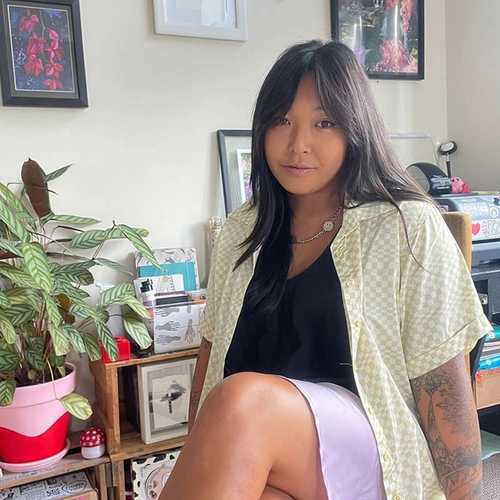 A woman sitting in front of a wall with framed art work. She has long black hair and is wearing a black t-shirt, pale yellow shirt, and purple skirt. 