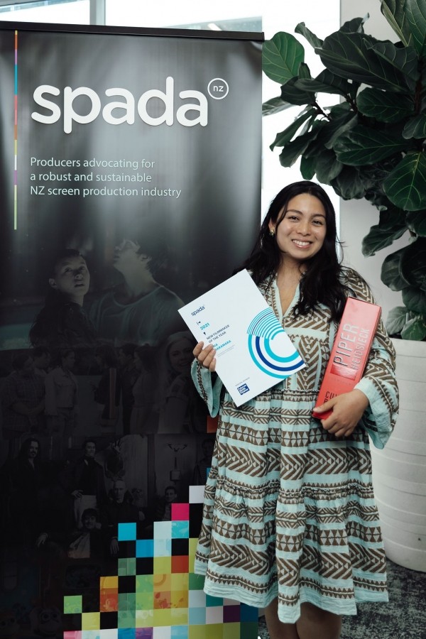 A Filipina in a blue and brown patterned dress smiling while holding a rectangular white document that says 'SPADA New Filmaker of the Year Award'