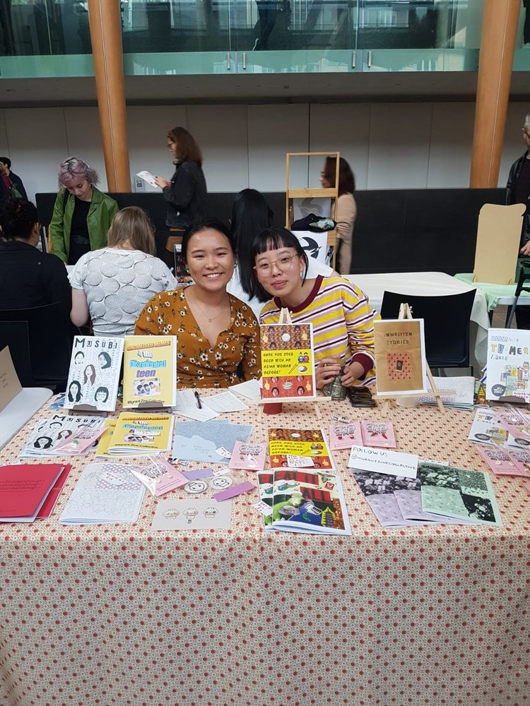 Two people smiling while manning a table displaying various colourful zines. 