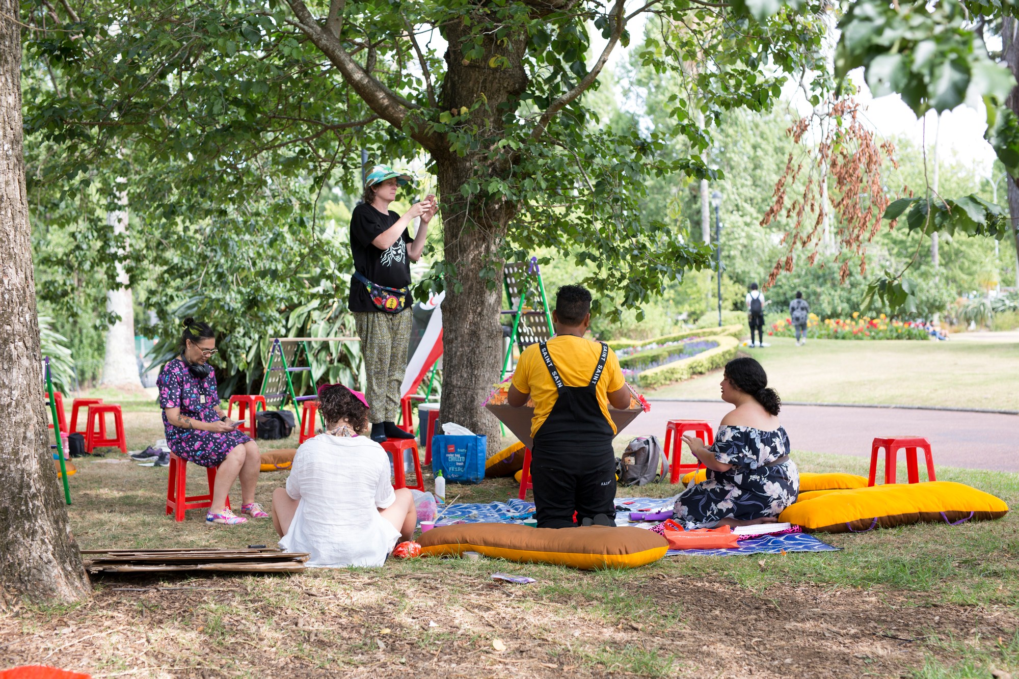 People sit and stand around a set up on the grass made up of colourful cushions, mats and plastic stools.