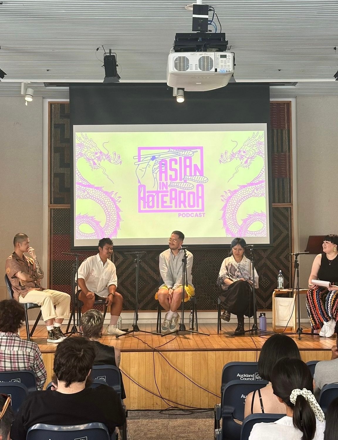 A stage with five people sitting on chairs in a panel discussion format. Behind, on a large screen is the text 'Asian in Aotearoa'