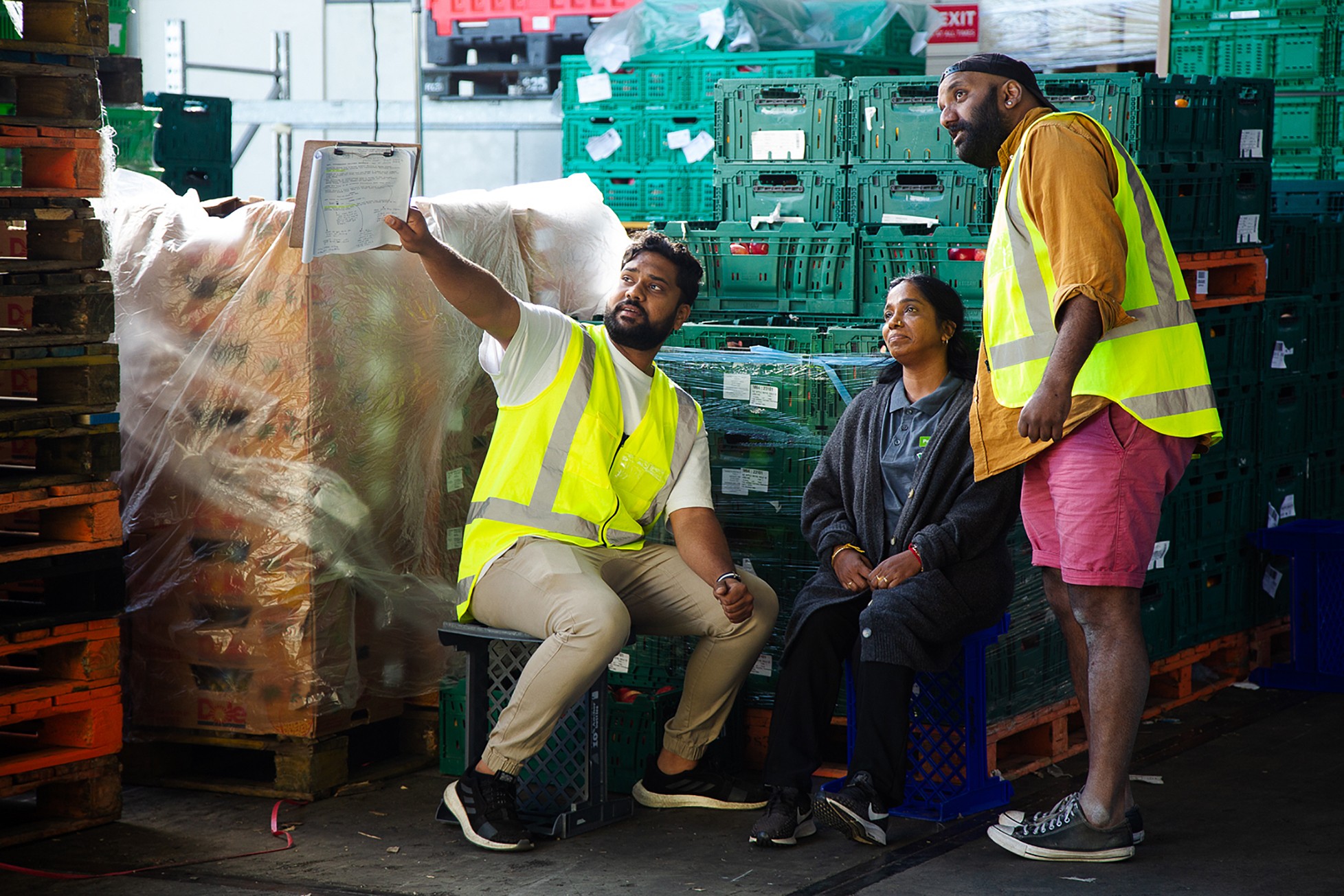 A woman in black pants and a dark gray cardigan sits on a plastic crate in between two men in high-visibility vests. The man on the left is holding a clipboard and is pointing into the distance.