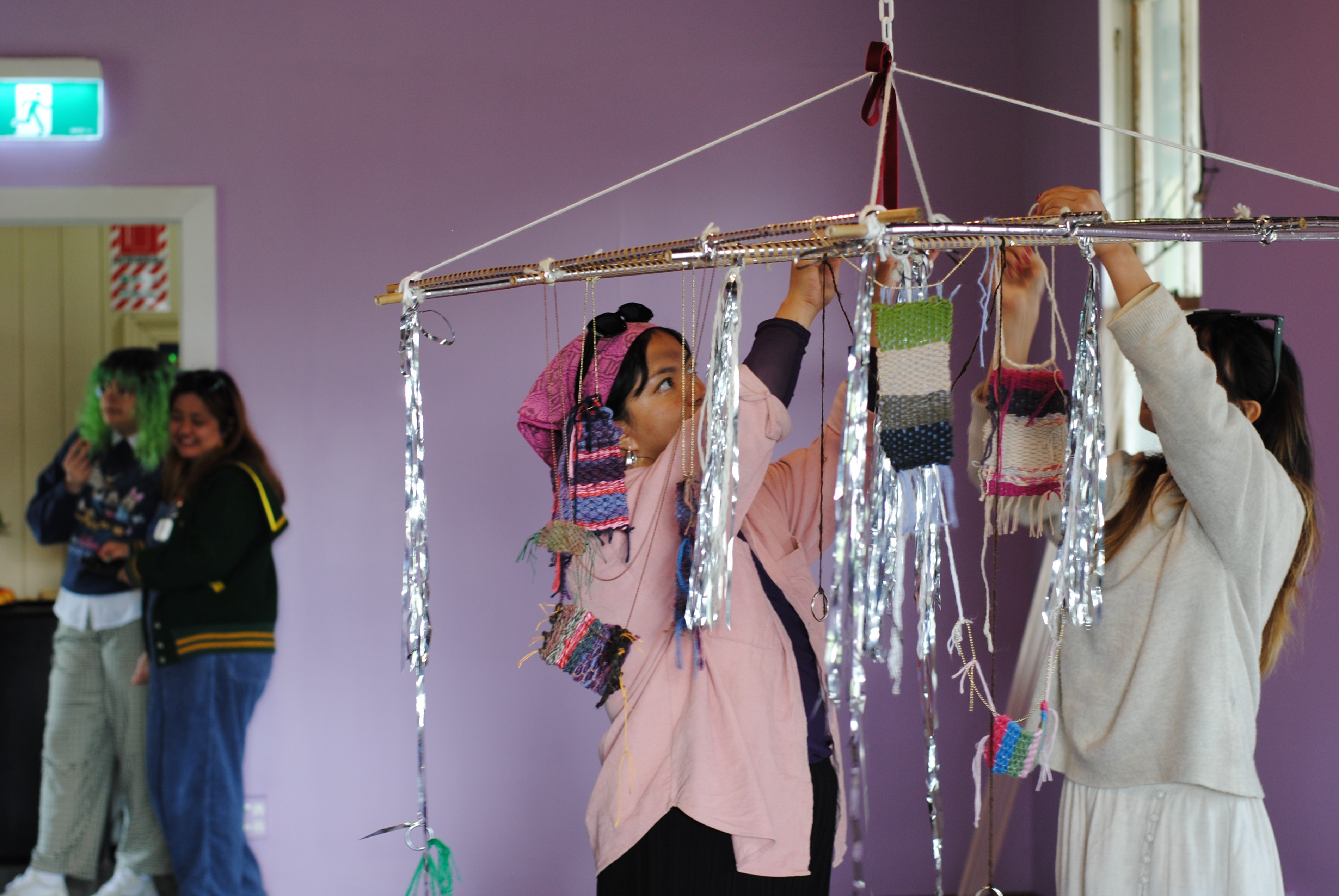 A person of Filipino descent wearing a pink bandanna and shirt with their friend in an all white outfit hanging small colourful weaving on a bamboo grid adorned with silver tape and silver tassels