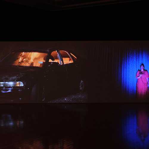An illuminated car in a dark art gallery, next to a video work showing a woman in a pink dress singing.