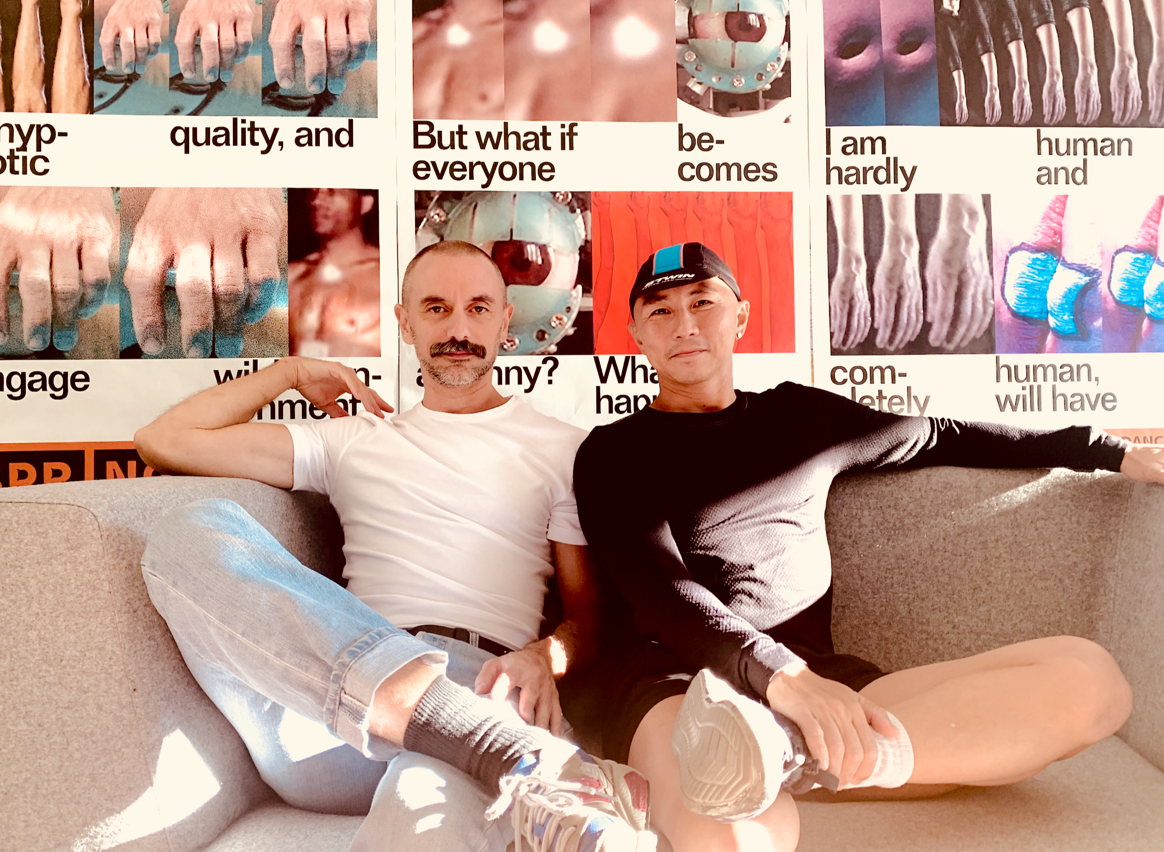 Two men sit on a couch together. The man on the left wears a white shirt and blue jeans and the other wears a cap and black shirt and shorts.