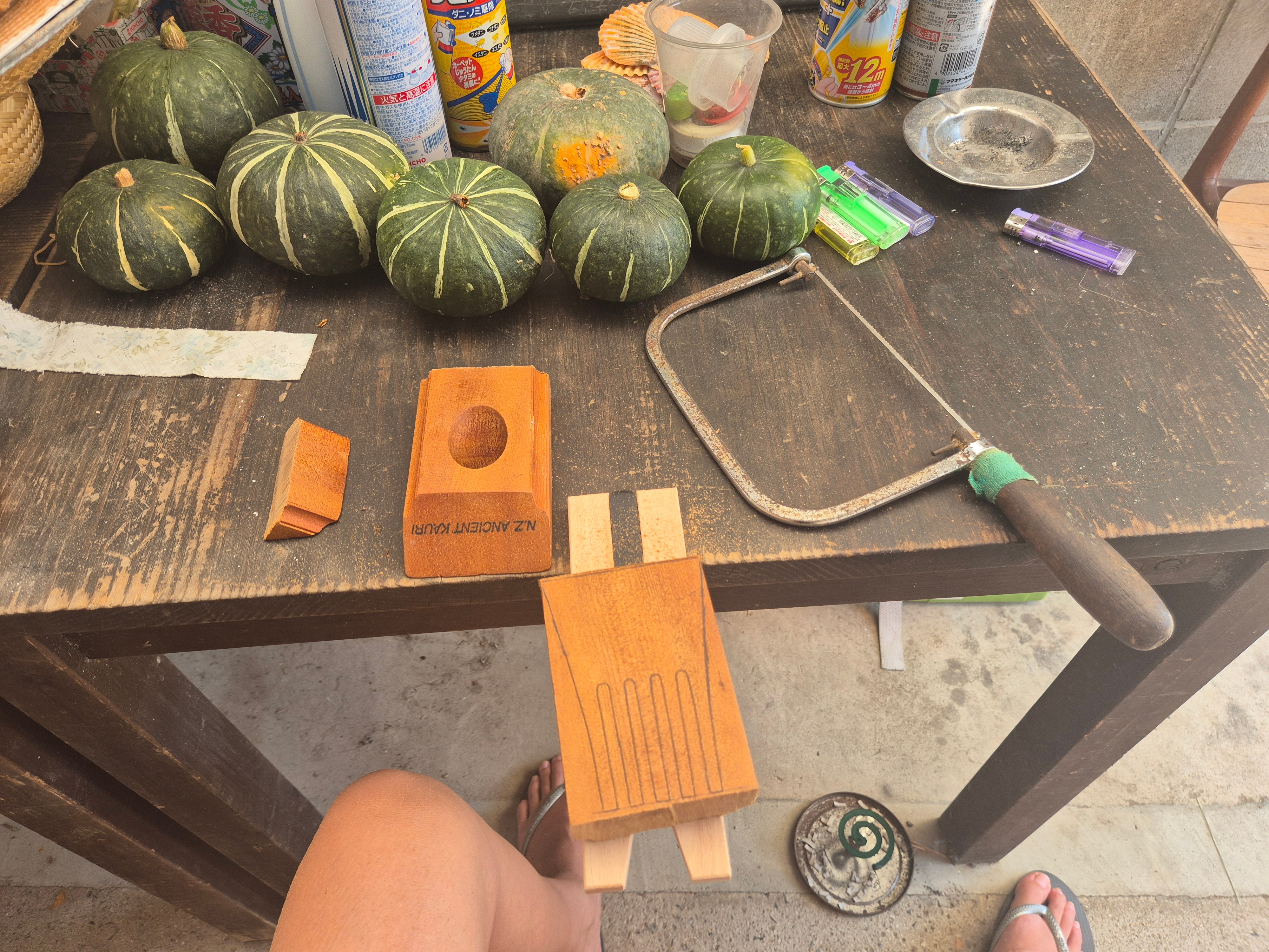 A person sitting down with woodwork in front of them. Spray cans, pumpkins and ephemera are scattered on the bench