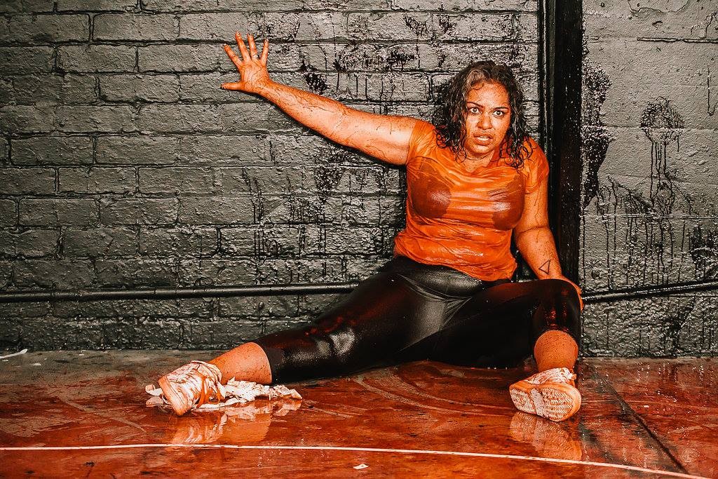 A woman leaning against a black brick wall covered in blood, with the floor also covered in blood. She is staring blankly into the audience