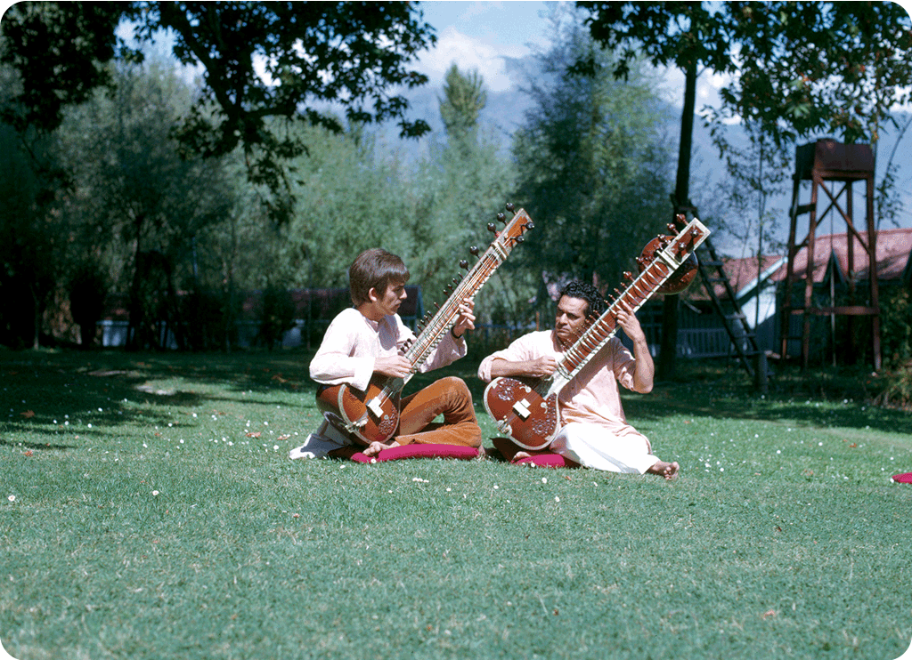 Harrison and Shankar sit on the grass playing sitar together.