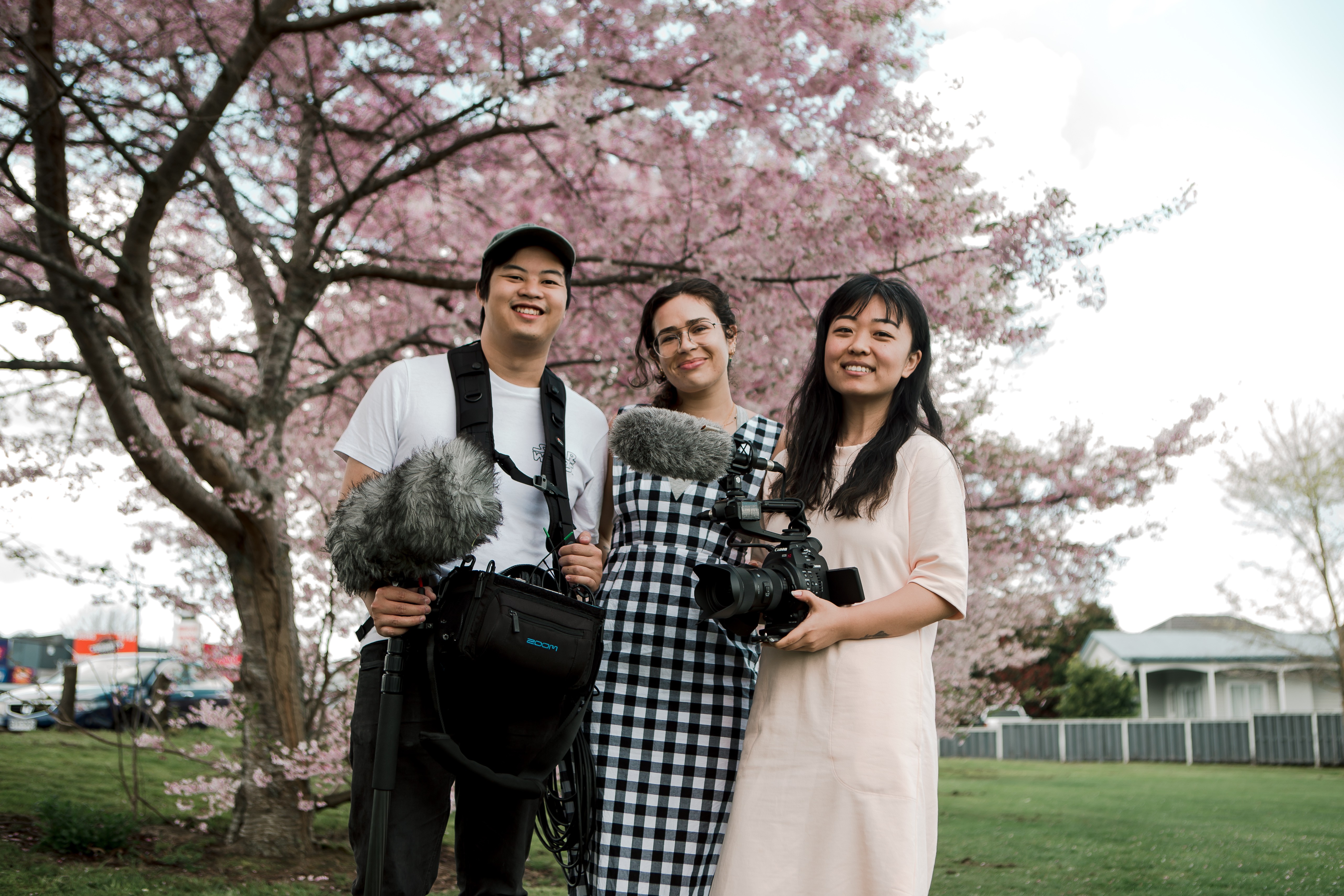 A man holding a boom,  a woman, and another woman holding a camera pose together in front of a blossoming tree.