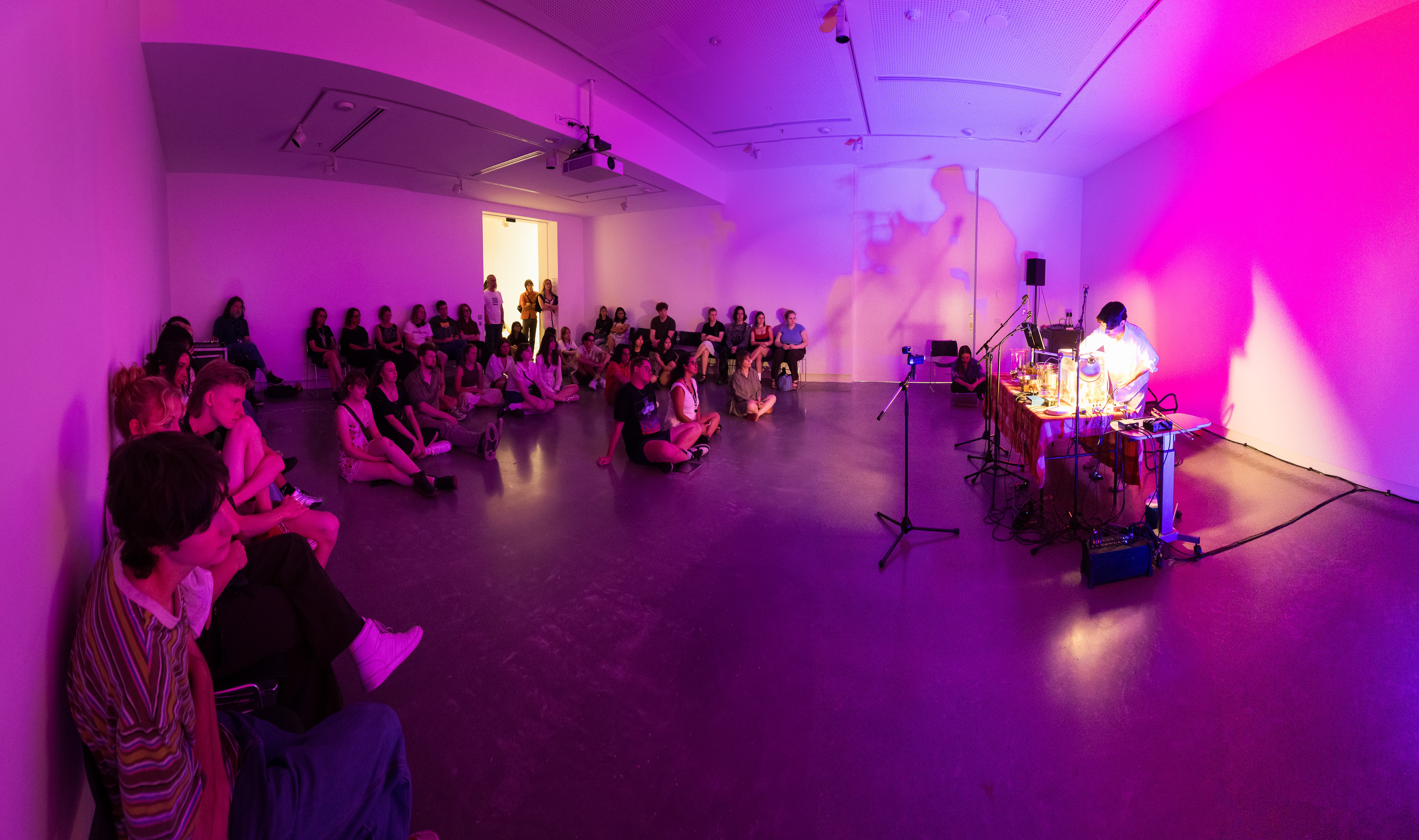 A room full of audience members sit, stand, watch and listen to the performer standing at a table of instruments, shrouded in purple light.