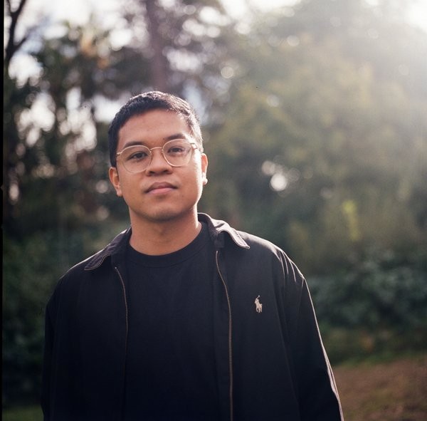 A man with glasses and a black jacket stands in front of some trees. Sunlight shines behind him.