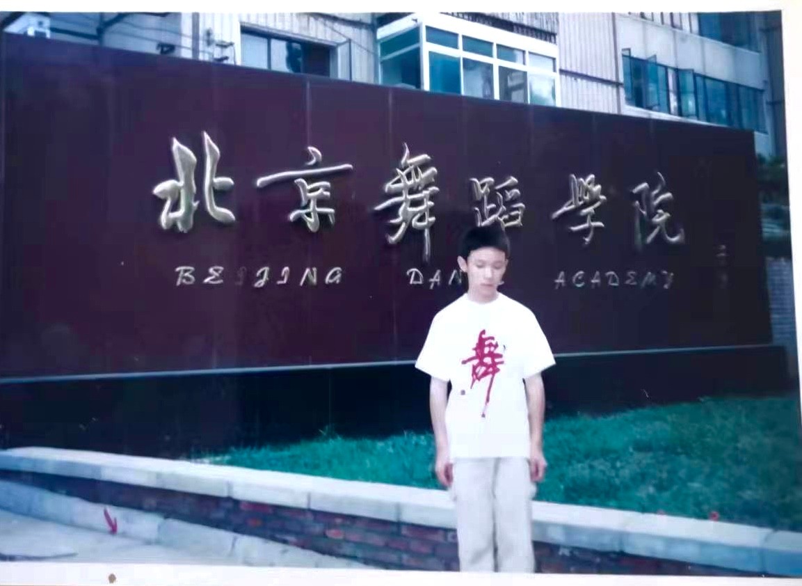 A young boy with a white shirt stands shyly in front of the Beijing Dance Academy sign